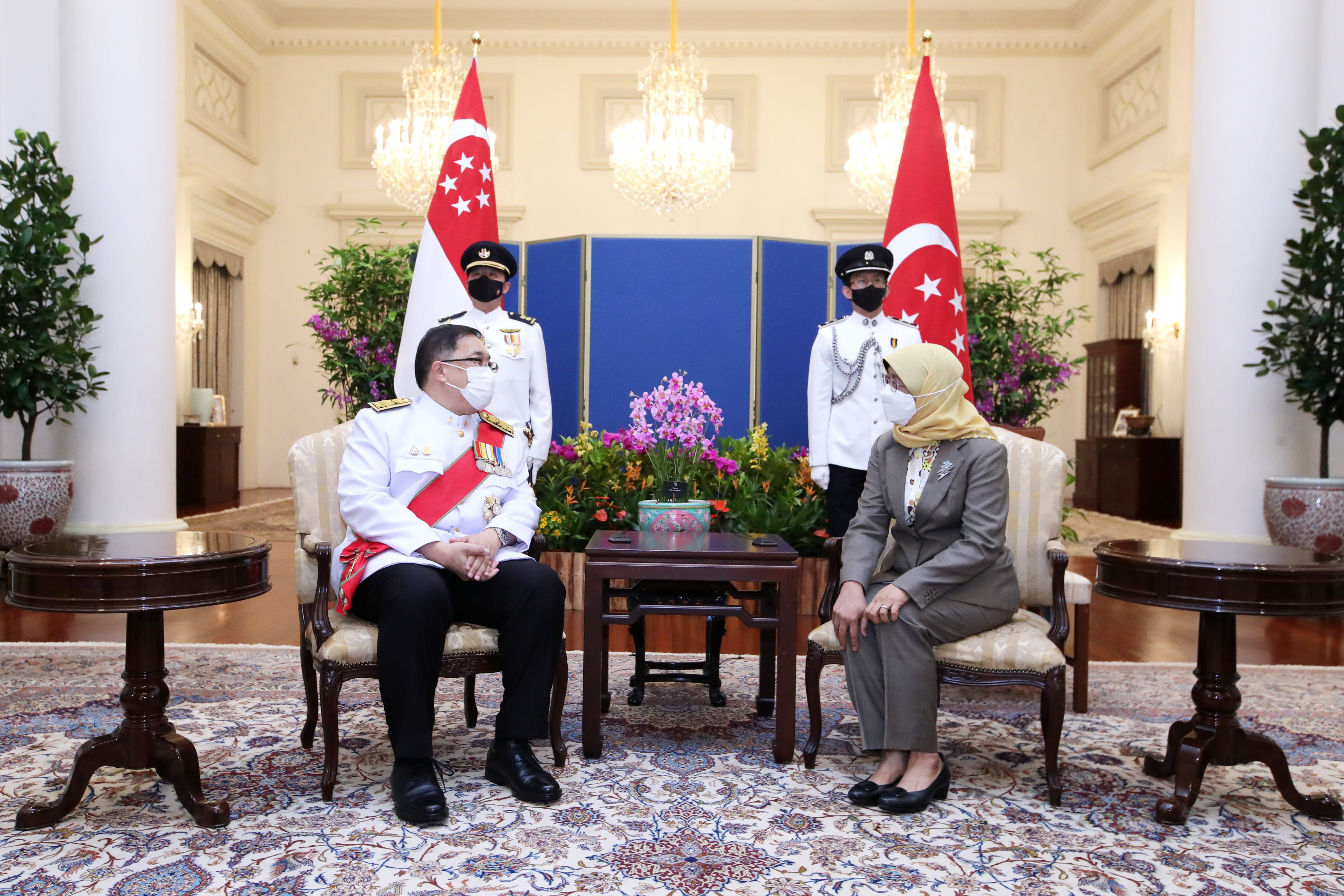 Two seated figures face each other, with Singapore flags and guards standing behind them.